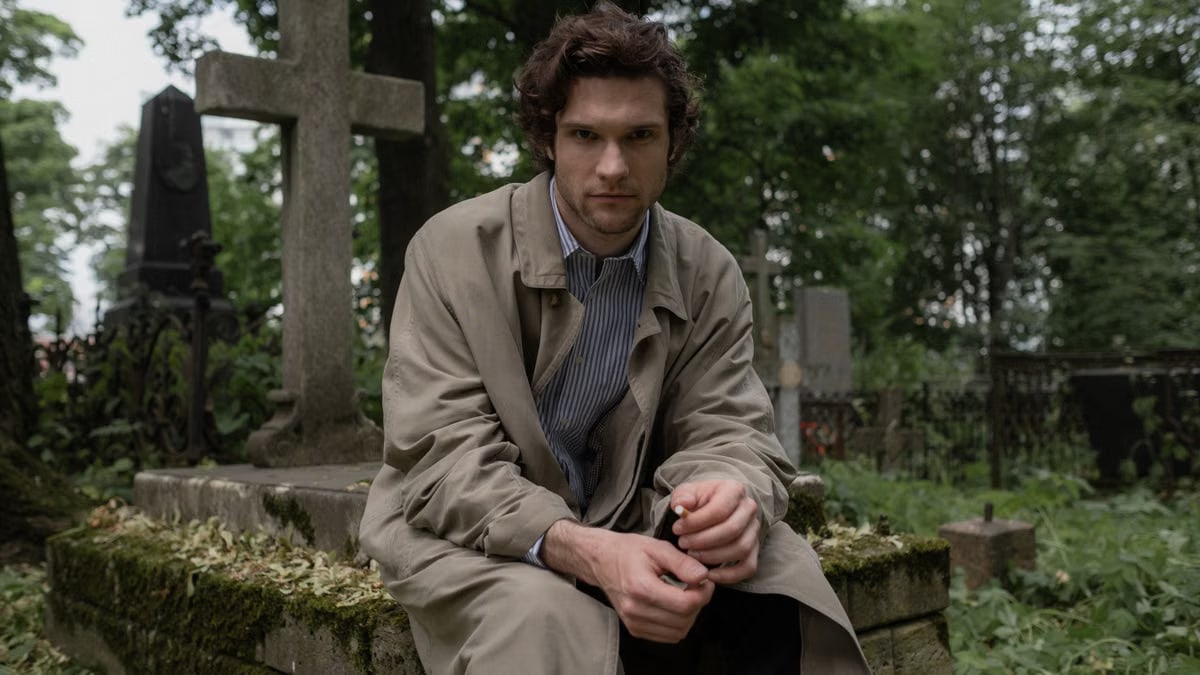 A man in a trench coat sits pensively near a headstone in a tranquil graveyard.